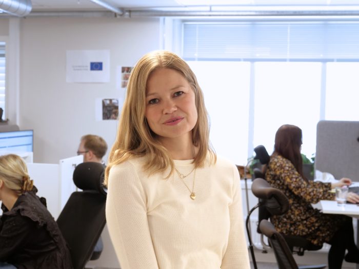 Kvinne med blondt hår og hvit genser står foran et kontorlandskap og ser alvorlig inn i kamera.