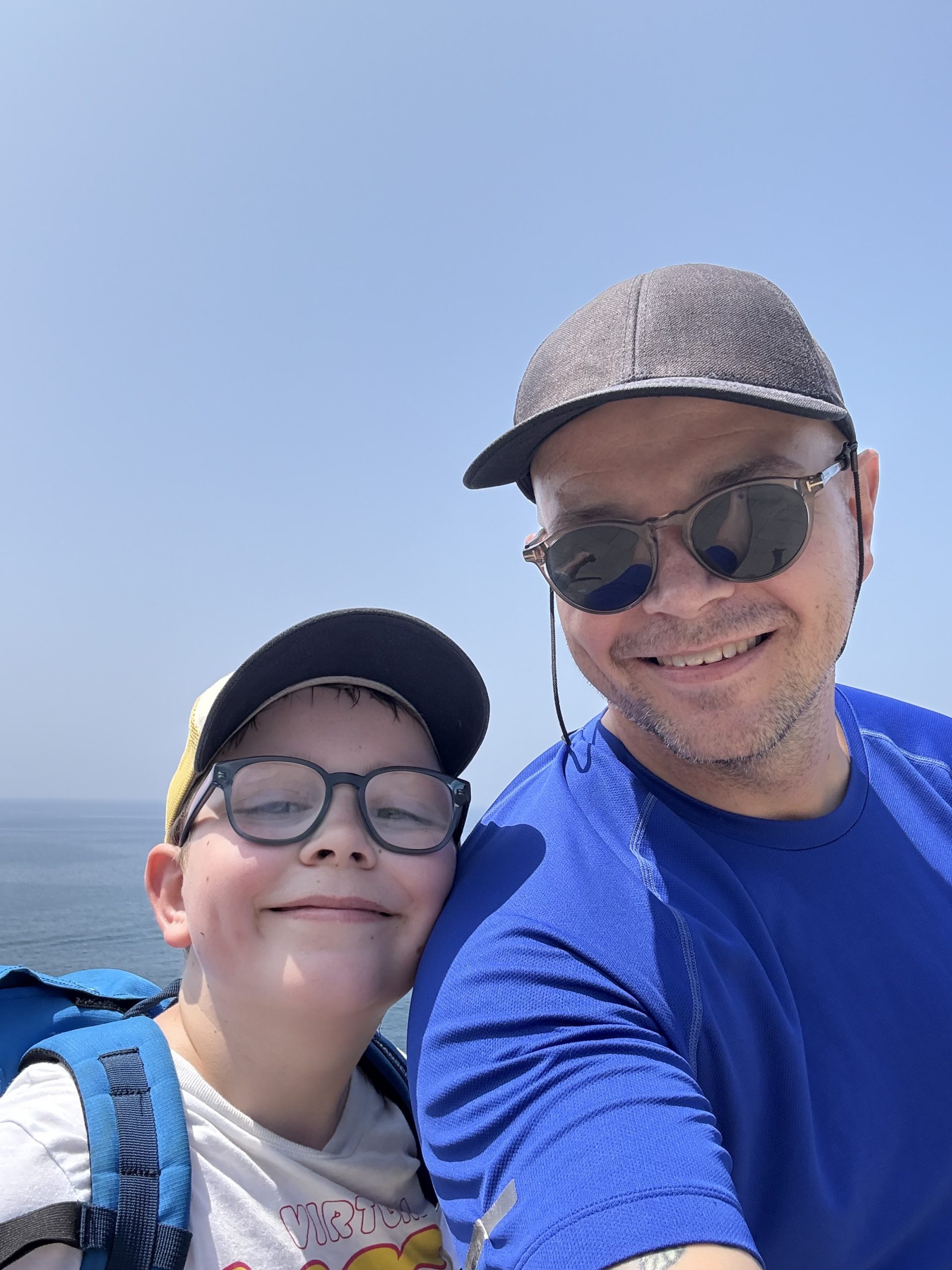 Småbarnsfar og sønn tar en selfie og ser smilende inn i kamera. Det er blå himmel og hav i bakgrunn.