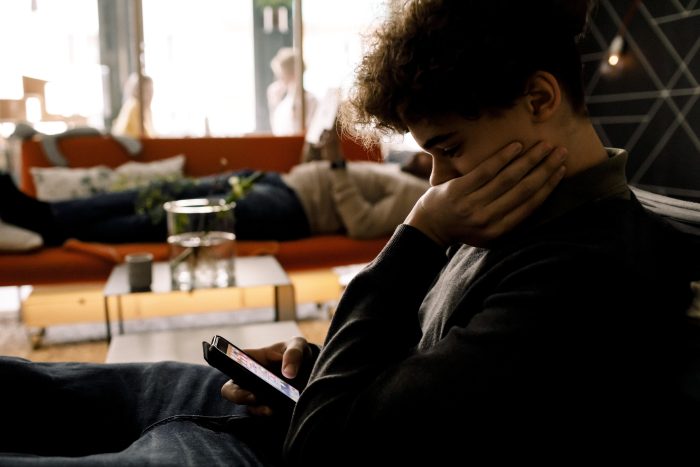 Gutt ser bekymret ned på mobilen sin med far i bakgrunn som ligger på en sofa