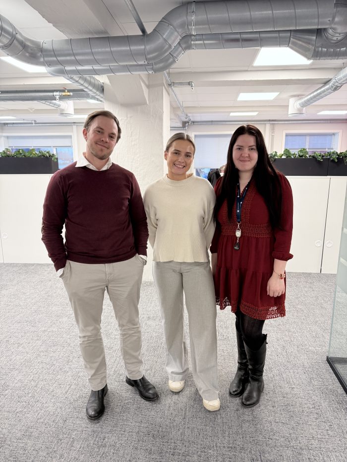 Tre unge ansatte, en mann og to kvinner, står i et åpent kontorlandskap og smiler mot kamera.