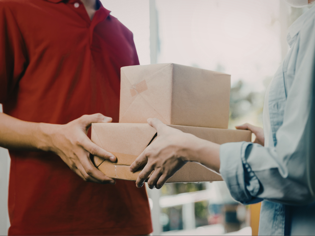 Nærbilde av postbud i rød skjorte som overrekker to bakker til en kunde kledd i en hvit skjorte.
